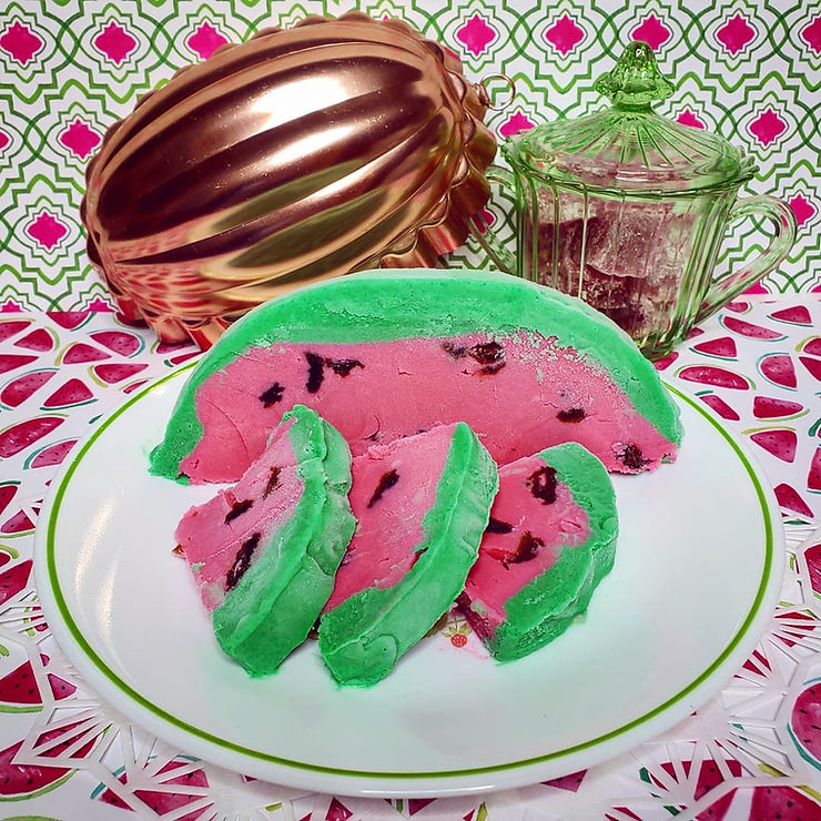 A frozen custard dessert mimicking a watermelon, plated in front of a copper 'melon' shaped mold and a green glass jar full of red candies.
